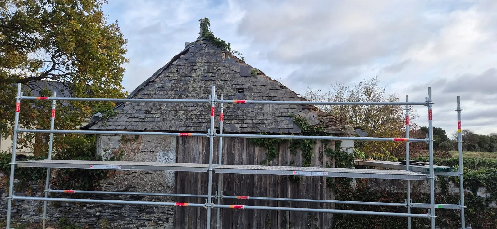 Grange en pierre à la vieille toiture en ardoise moussue, échafaudage monté avant réfection